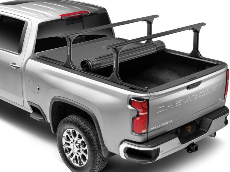 BAK Revolver X4TS hard roll-up tonneau cover rolled forward against the cab on a silver Chevrolet Silverado, with RealTruck crossbars mounted to the integrated T-slot rails, rear three-quarter angle on white background