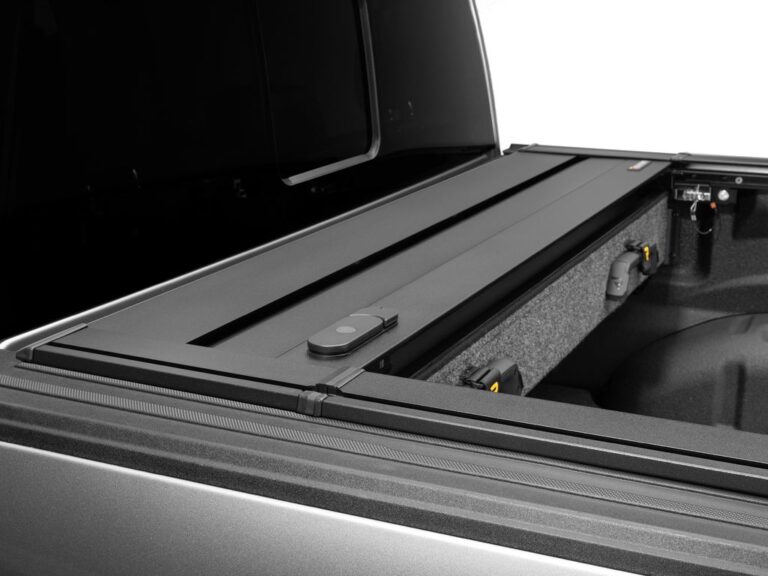 Close-up of the REALTRUCK branded badge on the tail-end of the closed RetraxPRO EZ-OFF FRC-4000 cover, showing the aluminum slat surface and matte black finish
