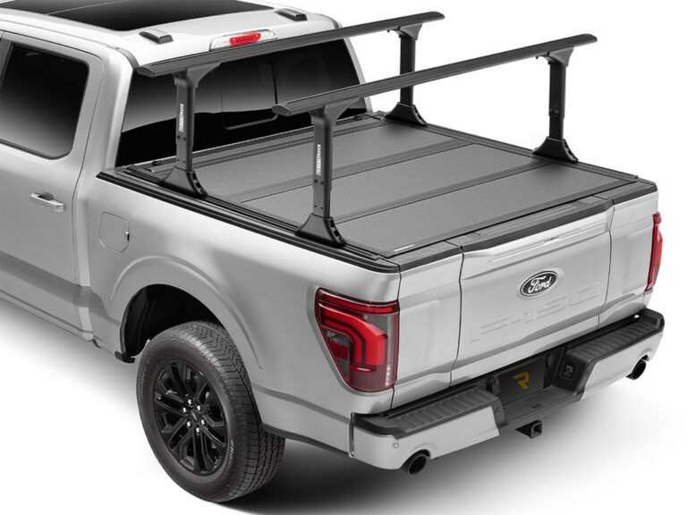 BAKFlip MX4 TS fully closed on a silver Ford F-150 with Yakima crossbars mounted on the T-slot rails, sitting flush with the bed rails, rear three-quarter angle on white background