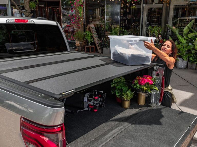 BAKFlip MX4 lifestyle image on a silver Ford F-150 with the cover open, woman loading a plastic storage tote into the bed in front of the Adorn Fine Flowers shop