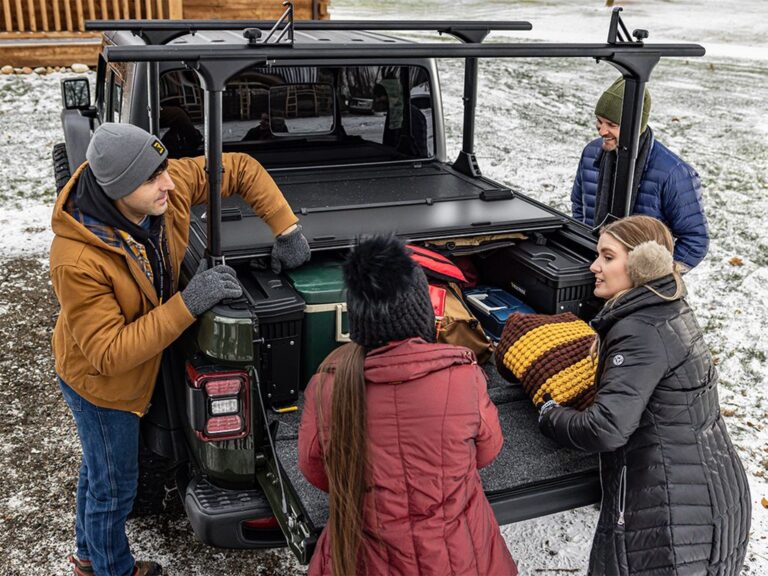 BAKFlip MX4 TS lifestyle shot on a dark green Jeep Gladiator in a snowy outdoor setting, cover fully open and folded against the cab, four people loading hard cases and outdoor gear into the bed