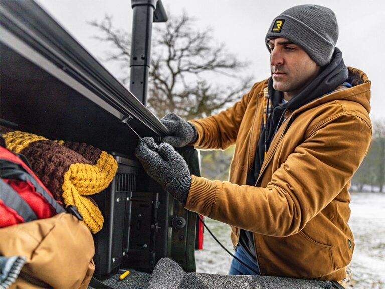 BAKFlip MX4 TS lifestyle close-up on a green Jeep Gladiator, hand reaching to the tailgate edge demonstrating the tight-fit clamp and weatherproof seal, snowy outdoor background