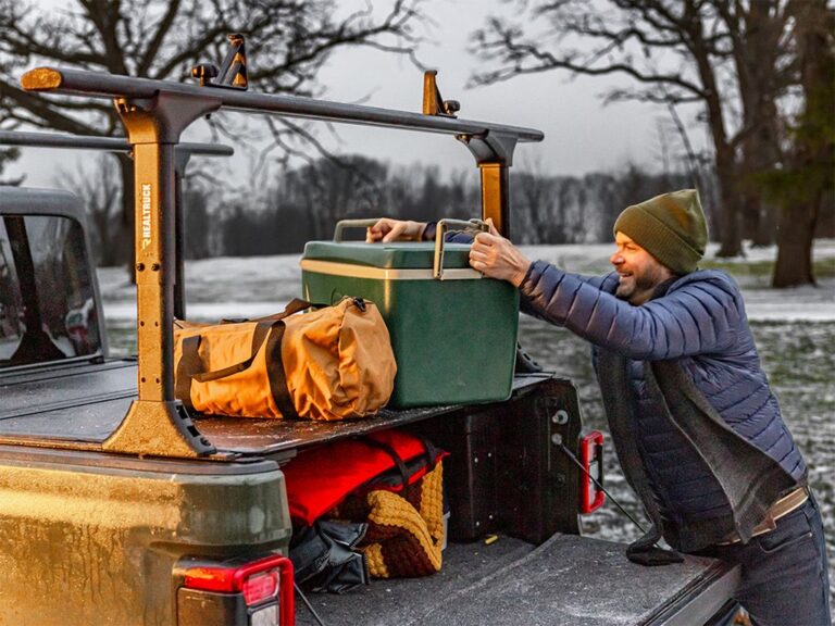 BAKFlip MX4 TS lifestyle image on a green Jeep Gladiator in a snowy setting, man in blue puffer jacket reaching up to load a green kayak onto the crossbars mounted on the T-slot rails