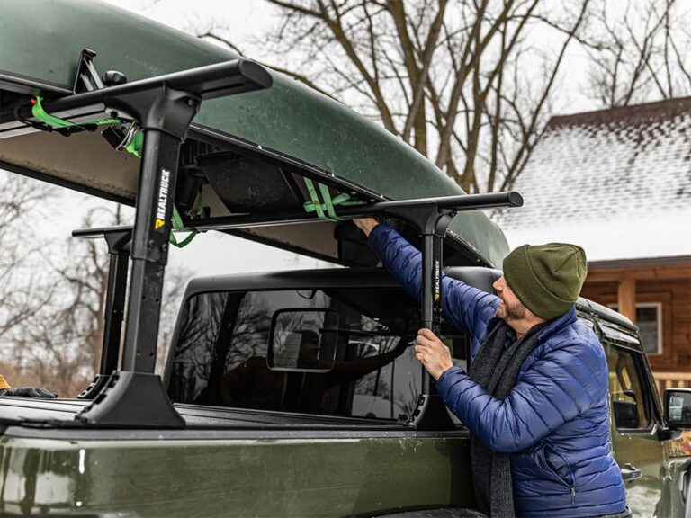 BAKFlip MX4 TS fully open and folded against the cab on a green Jeep Gladiator, man in blue puffer loading a green cooler and orange duffel into the bed, crossbars mounted above, snowy outdoor setting